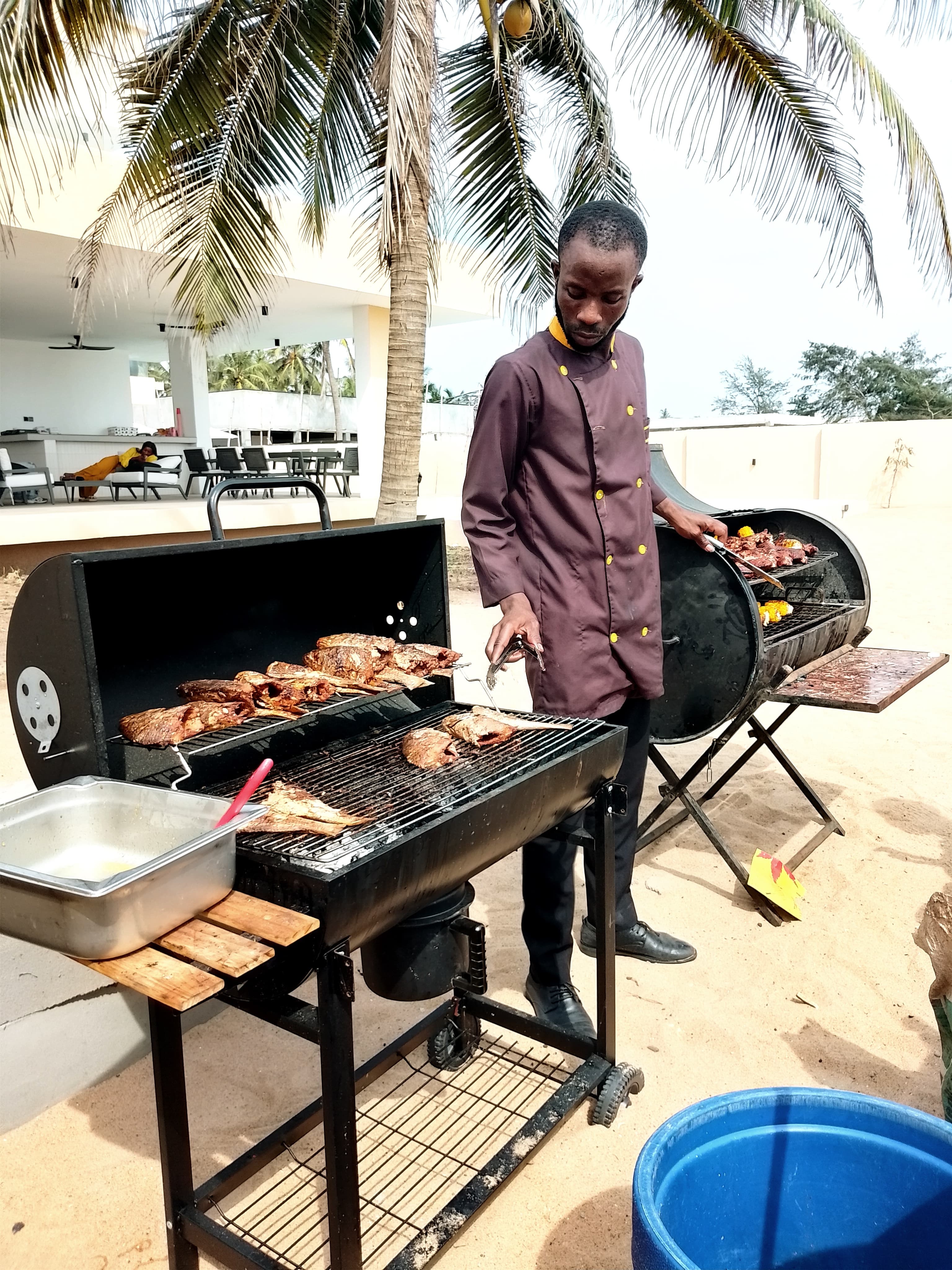 A chef grills whole fish and meat on large barrel grills under palm trees.
