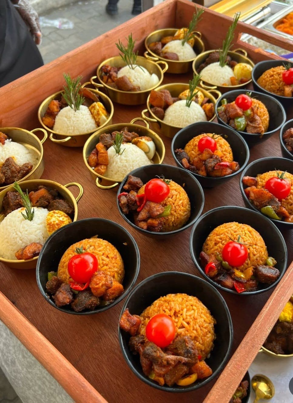 Small bowls of white and jollof rice with meat, rosemary, and tomatoes on a tray.