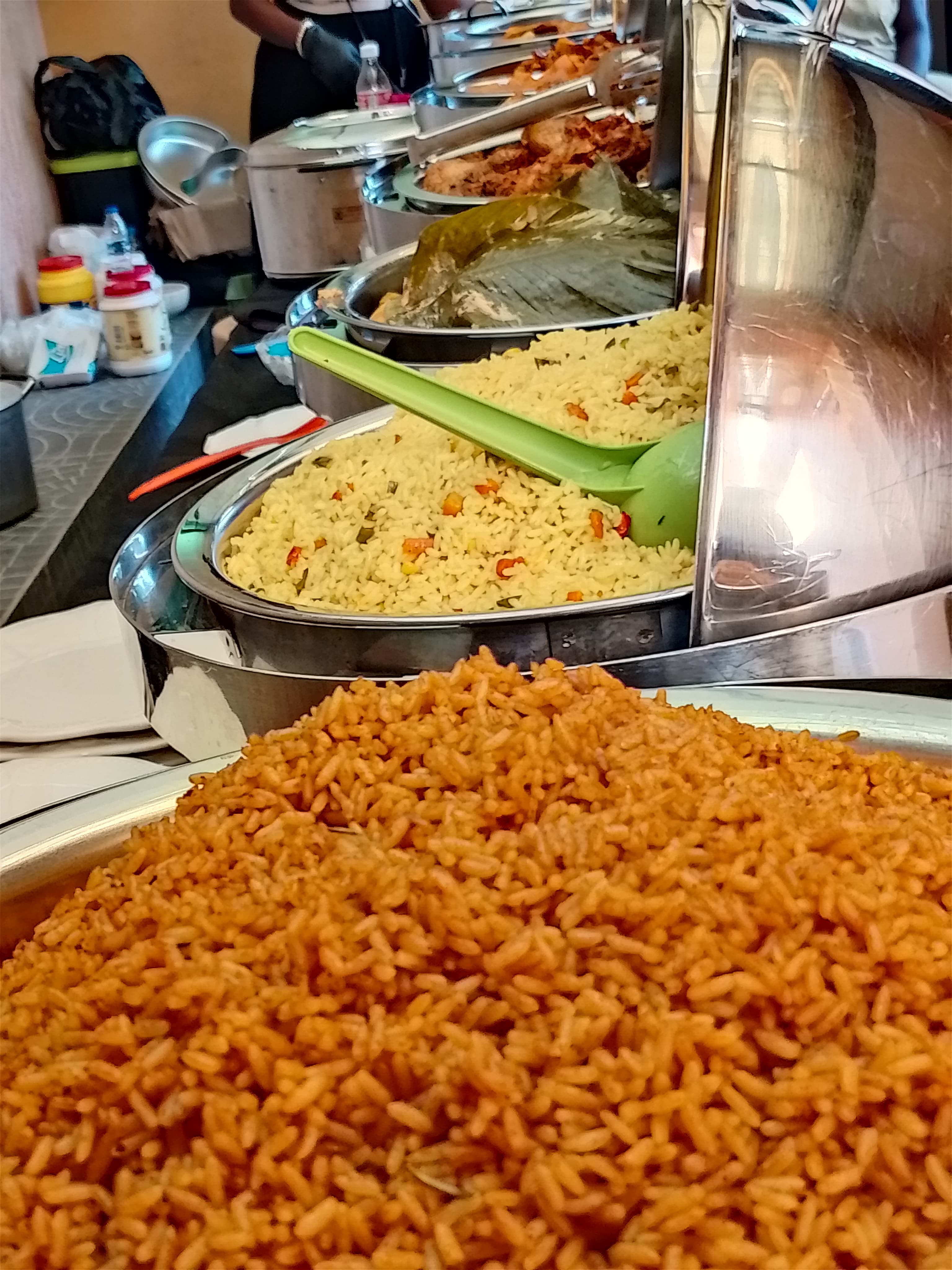 Buffet line featuring large metal trays of Jollof rice, fried rice, and fried chicken.